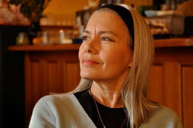 Candida Royalle, blonde hair and a black headband, thoughtful and smiling in a café